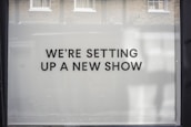 A storefront window features large, bold black text on a white background reading 'WE’RE SETTING UP A NEW SHOW'. The backdrop includes a reflection of brick walls and windows.