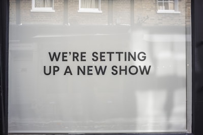 A storefront window features large, bold black text on a white background reading 'WE’RE SETTING UP A NEW SHOW'. The backdrop includes a reflection of brick walls and windows.