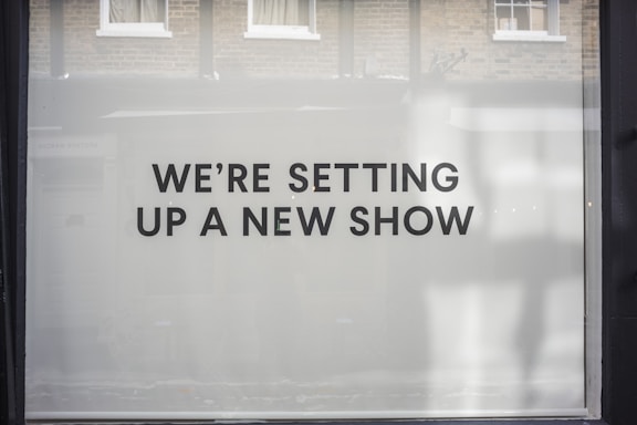 A storefront window features large, bold black text on a white background reading 'WE’RE SETTING UP A NEW SHOW'. The backdrop includes a reflection of brick walls and windows.