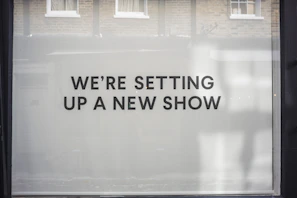 A storefront window features large, bold black text on a white background reading 'WE’RE SETTING UP A NEW SHOW'. The backdrop includes a reflection of brick walls and windows.