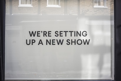 A storefront window features large, bold black text on a white background reading 'WE&rsquo;RE SETTING UP A NEW SHOW'. The backdrop includes a reflection of brick walls and windows.