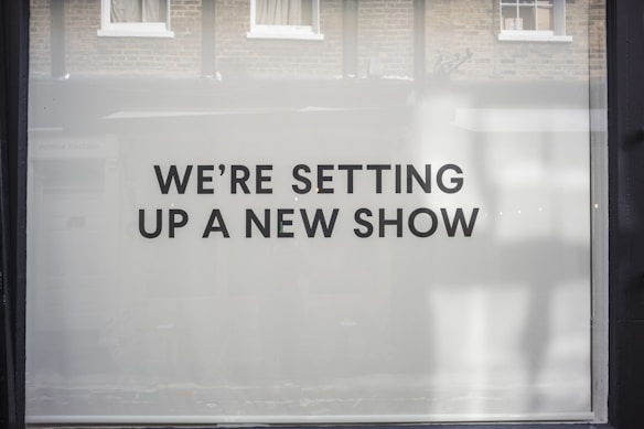 A storefront window features large, bold black text on a white background reading 'WE’RE SETTING UP A NEW SHOW'. The backdrop includes a reflection of brick walls and windows.