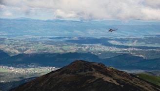 A helicopter flies over a mountainous landscape with vast open fields and scattered villages visible below. The sky is partly cloudy, casting shadows over the terrain. The scene offers a panoramic view of distant mountain ranges and lush greenery.