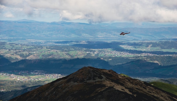 A helicopter flies over a mountainous landscape with vast open fields and scattered villages visible below. The sky is partly cloudy, casting shadows over the terrain. The scene offers a panoramic view of distant mountain ranges and lush greenery.