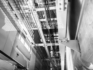 An interior view of an elevator shaft showing cables, metal beams, and structural components. The perspective is angular, focusing on the vertical lines and intricate machinery.