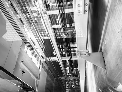 An interior view of an elevator shaft showing cables, metal beams, and structural components. The perspective is angular, focusing on the vertical lines and intricate machinery.