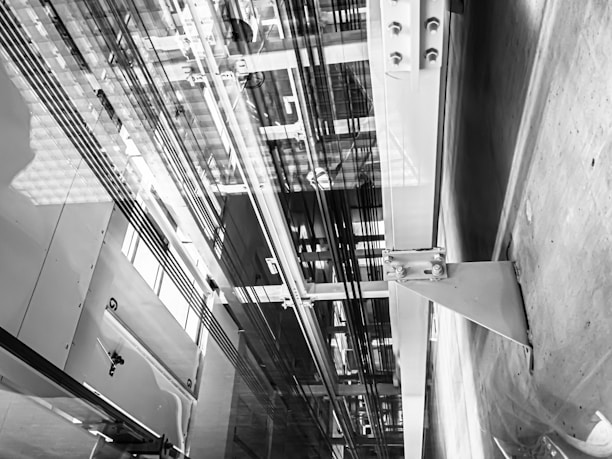 An interior view of an elevator shaft showing cables, metal beams, and structural components. The perspective is angular, focusing on the vertical lines and intricate machinery.