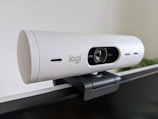 A white, speckled webcam mounted on the edge of a computer monitor. The lens is centered, with the brand's logo visible. The webcam has a cylindrical shape and is attached using a black mounting clip. The background is a neutral-colored wall with some greenery blurred in the corner.