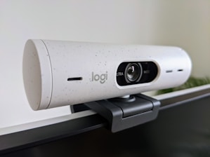 A white, speckled webcam mounted on the edge of a computer monitor. The lens is centered, with the brand's logo visible. The webcam has a cylindrical shape and is attached using a black mounting clip. The background is a neutral-colored wall with some greenery blurred in the corner.