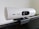 A white, speckled webcam mounted on the edge of a computer monitor. The lens is centered, with the brand's logo visible. The webcam has a cylindrical shape and is attached using a black mounting clip. The background is a neutral-colored wall with some greenery blurred in the corner.