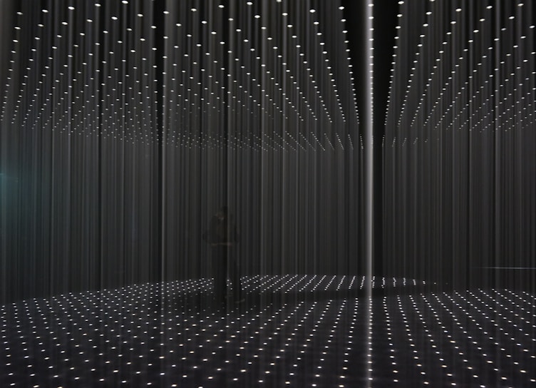 An installation art piece featuring a room filled with long strands of illuminated lights hanging from the ceiling, creating a grid-like pattern. The floor reflects the arranged lights, enhancing the three-dimensional effect. The space appears dark, with the lights providing the primary source of illumination.
