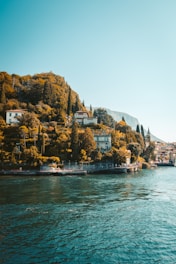 A serene lakeside scene with lush greenery and elegant villas nestled among tall cypress trees along the hillside. The calm waters of the lake reflect the blue sky above, creating a peaceful and tranquil atmosphere.