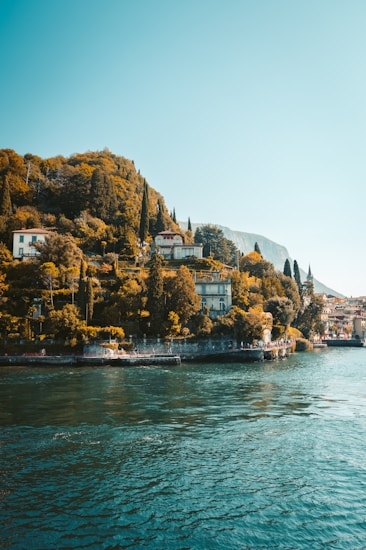 A serene lakeside scene with lush greenery and elegant villas nestled among tall cypress trees along the hillside. The calm waters of the lake reflect the blue sky above, creating a peaceful and tranquil atmosphere.