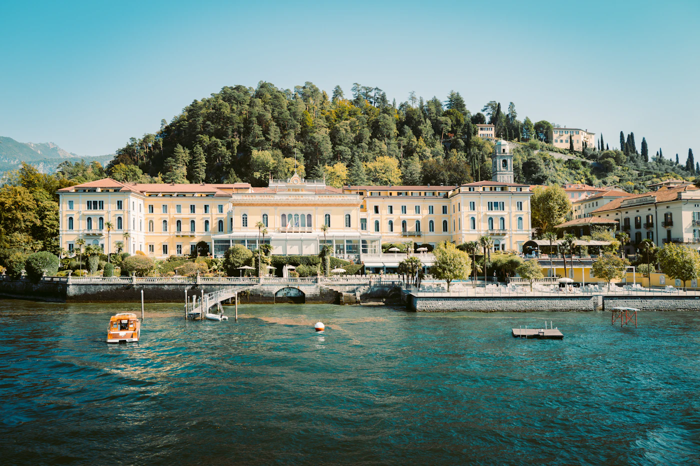Bellagio's waterfront seen from the lake, a long pale-yellow palace hotel below a wooded promontory