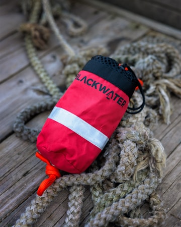 A red bag with a reflective silver stripe labeled 'BLACKWATER' rests on a wooden surface intertwined with thick, braided ropes. The bag features a black top with drawstring closure.