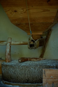 A rustic, manual grain mill with a large stone grinding wheel. Wooden components include a hopper above and a handle attached to the millstone. The surrounding area appears to be a wooden enclosure, giving an old-fashioned and traditional feel.