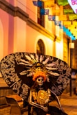 A person dressed in an elaborate costume featuring a large black feathered headpiece with intricate white designs, resembling traditional Mexican motifs. Their face is painted in a skull pattern, typical of Dia de los Muertos celebrations. The background shows a colorful street with vibrant papel picado banners hanging above.