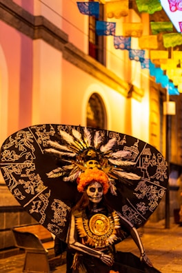 A person dressed in an elaborate costume featuring a large black feathered headpiece with intricate white designs, resembling traditional Mexican motifs. Their face is painted in a skull pattern, typical of Dia de los Muertos celebrations. The background shows a colorful street with vibrant papel picado banners hanging above.
