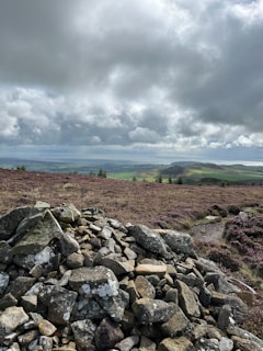 A pile of rocks is situated in the foreground, surrounded by a landscape of rolling heather-covered hills. The sky above is filled with thick, dramatic clouds, casting a muted light over the scene. In the distance, patches of green land can be seen, adding contrast to the earthy tones.
