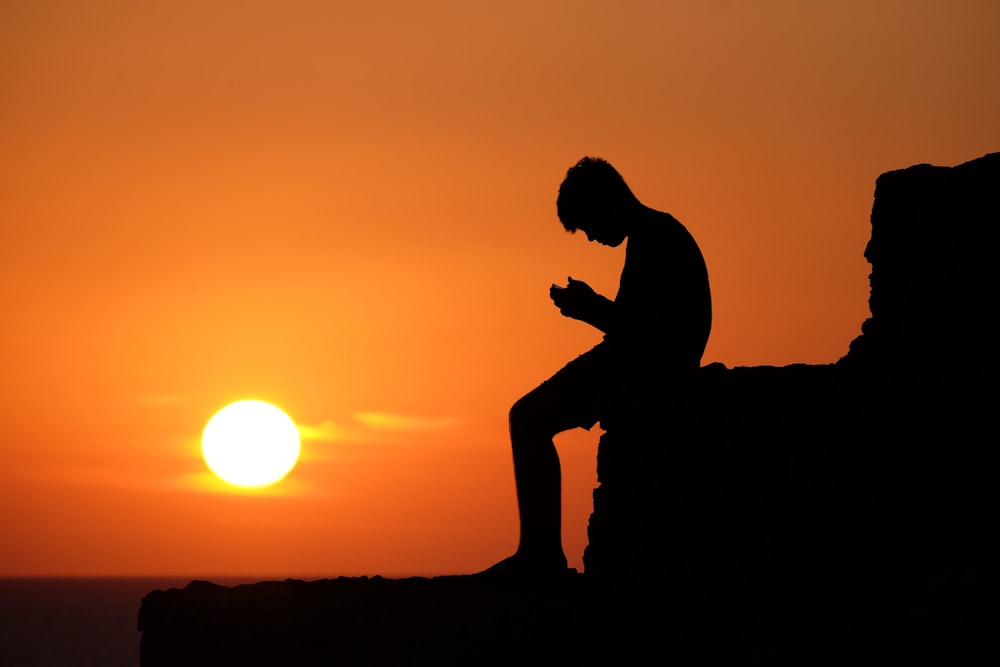 a silhouette of a man sitting on a rock at sunset