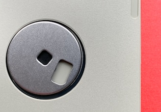 A close-up view of a vintage 3.5-inch floppy disk. The central metal shutter is prominently featured, showing a reflective metallic surface. The plastic casing has a textured light gray finish, and the edge of the disk is visible, revealing a vibrant red strip.