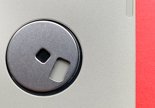 A close-up view of a vintage 3.5-inch floppy disk. The central metal shutter is prominently featured, showing a reflective metallic surface. The plastic casing has a textured light gray finish, and the edge of the disk is visible, revealing a vibrant red strip.