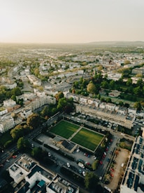 An aerial view of a cityscape featuring a mix of residential and commercial buildings. Lush green parks and squares are interspersed between the structures. Roads are lined with parked vehicles, and the layout suggests a well-organized urban area. The horizon shows a gradual transition into distant landscapes under a clear sky.