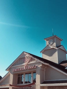 A building with a clock tower featuring a large sign that reads 'Boston Pizza' on the facade. The architecture includes a steeply pitched roof and large windows, set against a clear blue sky.