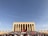 A large crowd of people gathers in front of a monumental building adorned with a massive red flag featuring a star and crescent. The structure is characterized by its tall pillars and grand architectural style, surrounded by a clear blue sky.