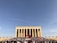 A large crowd of people gathers in front of a monumental building adorned with a massive red flag featuring a star and crescent. The structure is characterized by its tall pillars and grand architectural style, surrounded by a clear blue sky.