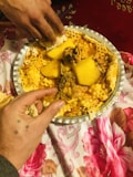 A platter of food containing pieces of meat, large chunks of potato, and a wet, porous bread-like substance. Two hands are reaching into the dish, suggesting it is meant to be shared communally. The platter is placed on a floral patterned tablecloth.