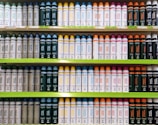 Several shelves in a store displaying neatly organized bottles of various personal care products. The bottles come in different colors and are arranged by type and purpose, with labels clearly visible. The shelves are green, and the organization gives a sense of order and cleanliness.