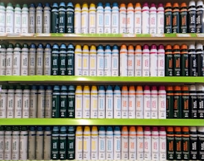 Several shelves in a store displaying neatly organized bottles of various personal care products. The bottles come in different colors and are arranged by type and purpose, with labels clearly visible. The shelves are green, and the organization gives a sense of order and cleanliness.