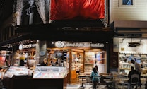An evening scene of a charming street cafe with a warm ambiance. The storefront is well-lit, showcasing displays of sweets and desserts in glass cases. People are seated outside, enjoying the atmosphere. The cafe has decorative lights and a large red banner above it.