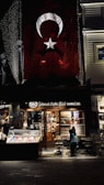 There is a large Turkish flag hanging on a building. Below the flag, there is a brightly lit café named 'Şehzade Cafe Erol' with a storefront showcasing various items. Outside, a few people are seated at tables. The setting appears to be in an urban area, possibly a street at night with some decorative lights.