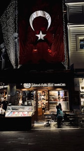 There is a large Turkish flag hanging on a building. Below the flag, there is a brightly lit café named 'Şehzade Cafe Erol' with a storefront showcasing various items. Outside, a few people are seated at tables. The setting appears to be in an urban area, possibly a street at night with some decorative lights.