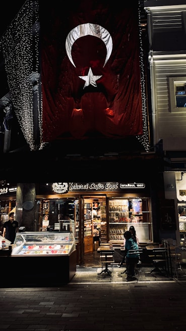 There is a large Turkish flag hanging on a building. Below the flag, there is a brightly lit café named 'Şehzade Cafe Erol' with a storefront showcasing various items. Outside, a few people are seated at tables. The setting appears to be in an urban area, possibly a street at night with some decorative lights.