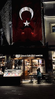 There is a large Turkish flag hanging on a building. Below the flag, there is a brightly lit caf&eacute; named 'Şehzade Cafe Erol' with a storefront showcasing various items. Outside, a few people are seated at tables. The setting appears to be in an urban area, possibly a street at night with some decorative lights.