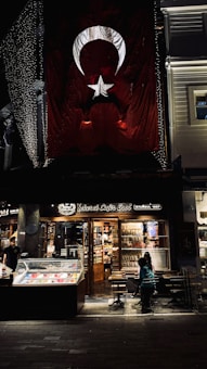 There is a large Turkish flag hanging on a building. Below the flag, there is a brightly lit caf&eacute; named 'Şehzade Cafe Erol' with a storefront showcasing various items. Outside, a few people are seated at tables. The setting appears to be in an urban area, possibly a street at night with some decorative lights.