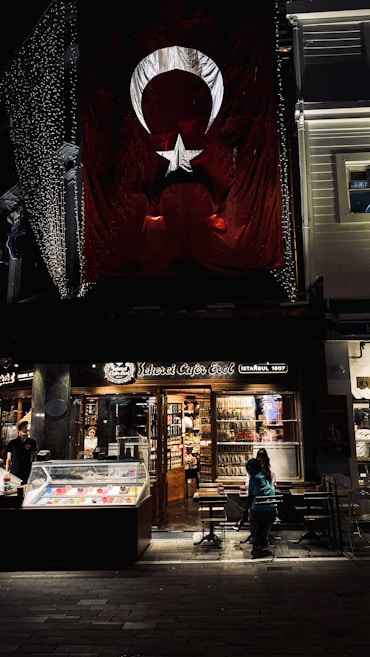 There is a large Turkish flag hanging on a building. Below the flag, there is a brightly lit caf&eacute; named 'Şehzade Cafe Erol' with a storefront showcasing various items. Outside, a few people are seated at tables. The setting appears to be in an urban area, possibly a street at night with some decorative lights.