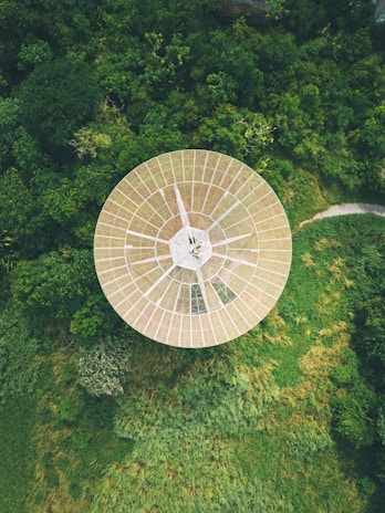 A large, circular satellite dish is nestled amidst dense green foliage, viewed from above. The structure is metallic with a grid-like pattern and contrasts with the vibrant greens surrounding it. The surrounding vegetation is lush and dense, suggesting a remote or natural setting.