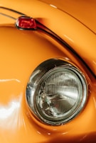 Close-up of a classic car's rounded headlight and orange body. The chrome ring around the headlight adds a vintage feel. The paint reflects light, showing a smooth and polished surface, with a small portion of a red taillight visible.