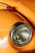 Close-up of a classic car's rounded headlight and orange body. The chrome ring around the headlight adds a vintage feel. The paint reflects light, showing a smooth and polished surface, with a small portion of a red taillight visible.