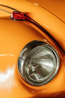 Close-up of a classic car's rounded headlight and orange body. The chrome ring around the headlight adds a vintage feel. The paint reflects light, showing a smooth and polished surface, with a small portion of a red taillight visible.