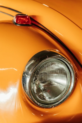 Close-up of a classic car's rounded headlight and orange body. The chrome ring around the headlight adds a vintage feel. The paint reflects light, showing a smooth and polished surface, with a small portion of a red taillight visible.
