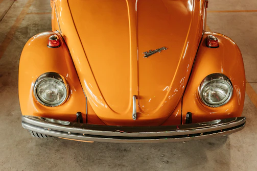 A classic orange car viewed from the front, featuring round headlights and a distinctive emblem on the hood. The vehicle is parked indoors, and the lighting highlights its shiny, smooth surface.