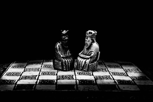 Two intricately detailed chess-like figures are positioned on a patterned, monochromatic chessboard. The scene is dramatic with deep shadows, focusing attention on the central figures in a dark, minimalist setting.