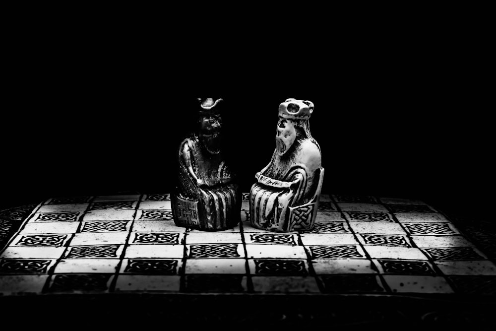 a black and white photo of two figurines sitting on a checkered table