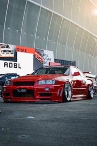 A custom red sports car with a sleek, aerodynamic design is parked in front of a large display with automotive-themed advertisements. The car features polished rims and a low suspension, indicating modifications for performance or aesthetic enhancement. The setting appears to be an automotive show or exhibition, with other cars partially visible in the background.