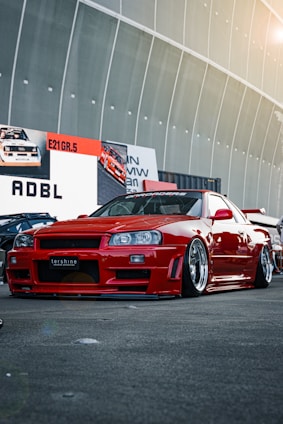 A custom red sports car with a sleek, aerodynamic design is parked in front of a large display with automotive-themed advertisements. The car features polished rims and a low suspension, indicating modifications for performance or aesthetic enhancement. The setting appears to be an automotive show or exhibition, with other cars partially visible in the background.